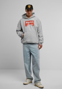 Grå hettegenser med "Market"-logo, frontlomme, brukt med lyseblå baggy jeans og beige sneakers. Svart caps med logo foran.