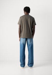 Mand med mørk hud og korte dreadlocks står med ryggen til, iført en mørkegrå t-shirt, løse blå jeans og hvide sko.