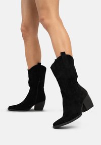 Botas de cowboy de ante negro con punta afilada, tacón cuadrado y costuras decorativas. Cuentan con una cremallera lateral para facilitar su uso.