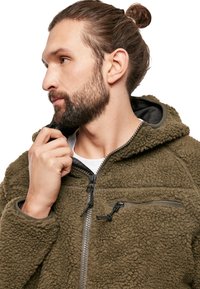 Veste en polaire vert olive avec fermeture éclair, extérieur texturé et capuche. Comprend une poche poitrine zippée et des cordons de serrage réglables.