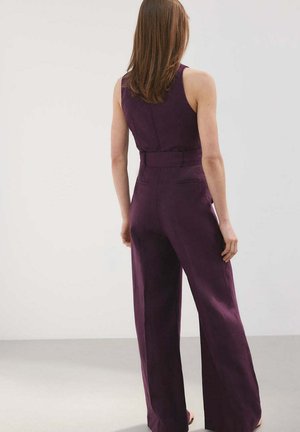 Mujer con cabello castaño con un mono sin mangas, de pierna ancha y cintura alta en color púrpura oscuro, de pie contra un fondo claro liso, de espaldas.