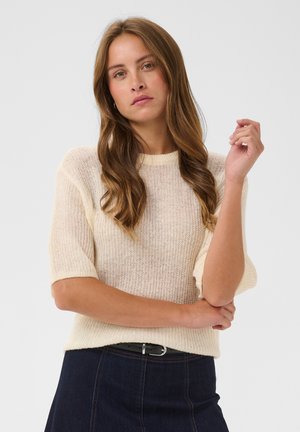 Femme aux longs cheveux bruns ondulés portant un pull léger beige à manches courtes en tricot et une jupe bleu foncé avec ceinture noire sur fond blanc.