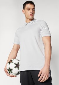 Svetlošedá polokošeľa s golierom a tromi gombíkmi, s bielym logom adidas na ľavom rukáve. Čierne šortky. Drží vzorovanú futbalovú loptu.