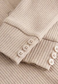 Ribbade beige tyget med en mjuk textur har tre ljusa knappar vid ärmsluten, vilket framhäver detaljerad sömnad och en figursydd design.