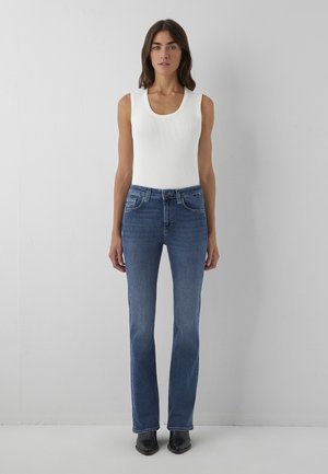 Woman with long dark hair wears sleeveless white top, blue straight-leg jeans, and black ankle boots, standing against a plain gray wall.