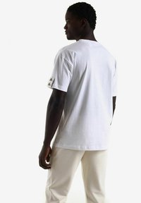 Camiseta blanca de algodón con cuello redondo y mangas cortas, que presenta un pequeño detalle de logo en la manga izquierda. Diseño minimalista.