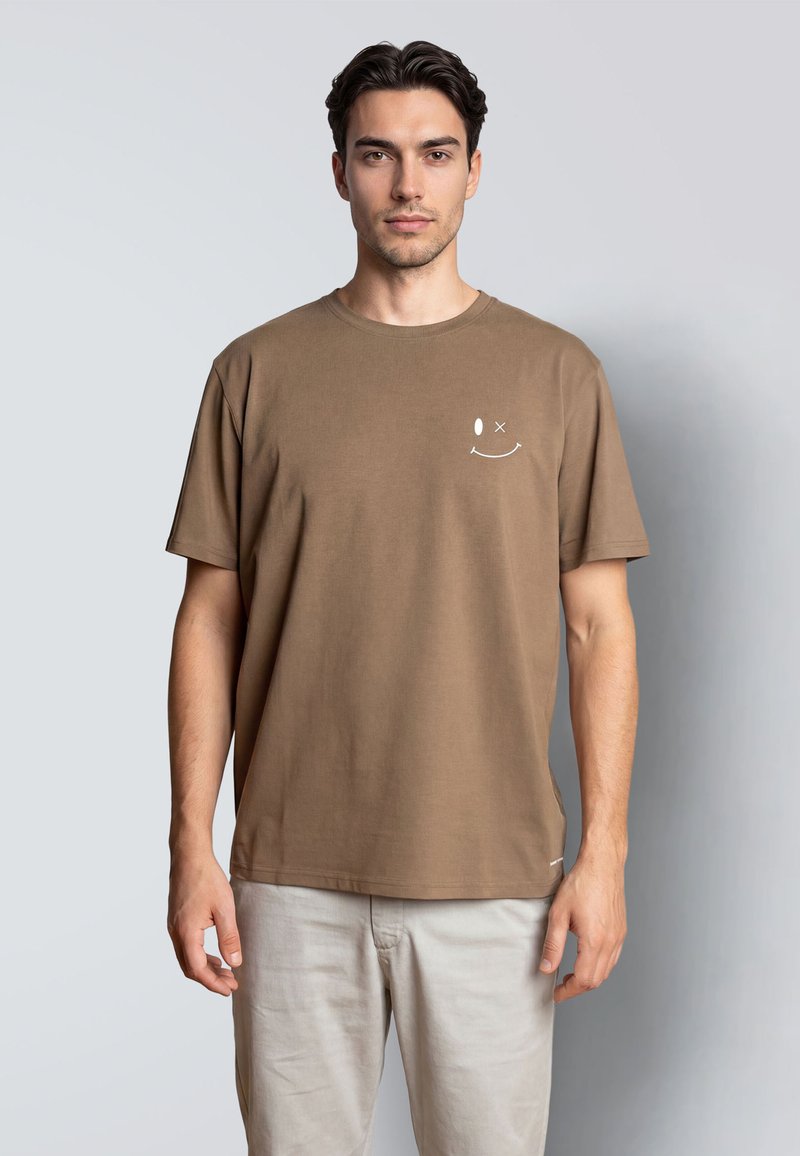 Joven con cabello oscuro que lleva una camiseta marrón con una pequeña carita sonriente guiñando un ojo en el pecho y pantalones beige claros.