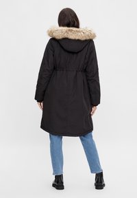 Czarna parka z kapturem wyściełanym futrem, taliowana, z długimi rękawami i lekko zaokrąglonym dołem, zestawiona z jasnoniebieskimi dżinsami i czarnymi butami.