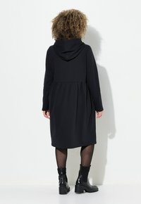 Schwarzes Kapuzenkleid mit ausgestellt geschnittenem Saum, aus weichem Stoff. Kombiniert mit klobigen schwarzen Knöchelstiefeln und strukturierten Strumpfhosen.