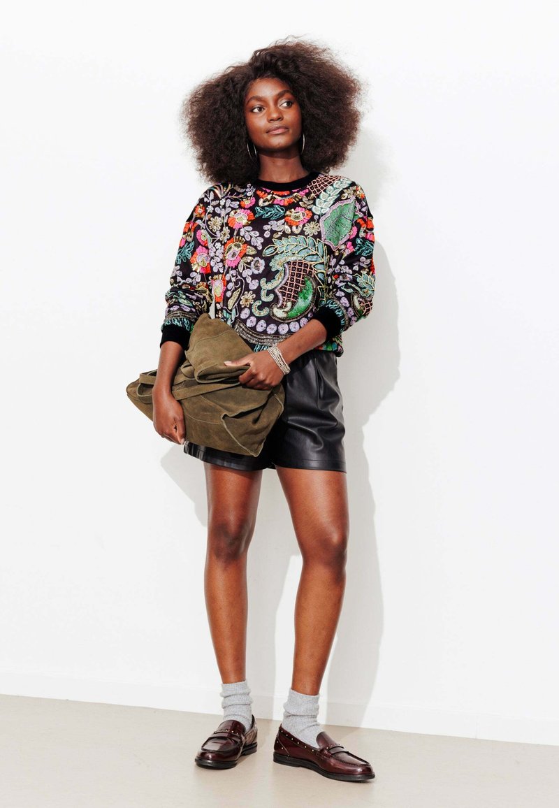 Pull noir brodé avec des motifs floraux, associé à un short en cuir noir. Tenant un sac en daim vert. Mocassins marron et chaussettes grises.