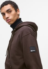 Rjava zip-up kapuca z teksturiranim zaključkom, črnisel ribast turtleneck in oznaka logotipa na rokavu. Ima kapuco z vrvico.