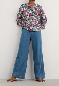 Person iført en farverig blomstret skjorte med lange ærmer, vidde blå jeans og beige remsandaler, stående mod en simpel beige væg.