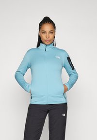 Kvinne med flettehår iført en lys blå zip-up jakke og svarte bukser, begge merket med The North Face-logoen, som står med hendene i lommene.