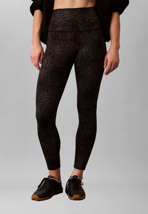 Persona con leggings negros de talle alto con un sutil estampado de leopardo y zapatillas negras con suela marrón, de pie sobre un fondo gris.