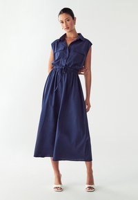 Abito senza maniche blu navy con design di camicia botton-up, due tasche sul petto e vita elastica. Il tessuto sembra leggero e liscio.