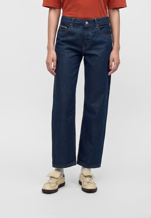 Personne portant un jean droit bleu foncé, des baskets beiges, des chaussettes blanches et une chemise orange sur un fond uni.