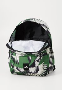 Green backpack with bird print, featuring a zippered opening, black interior, and a front pocket with a black logo patch.
