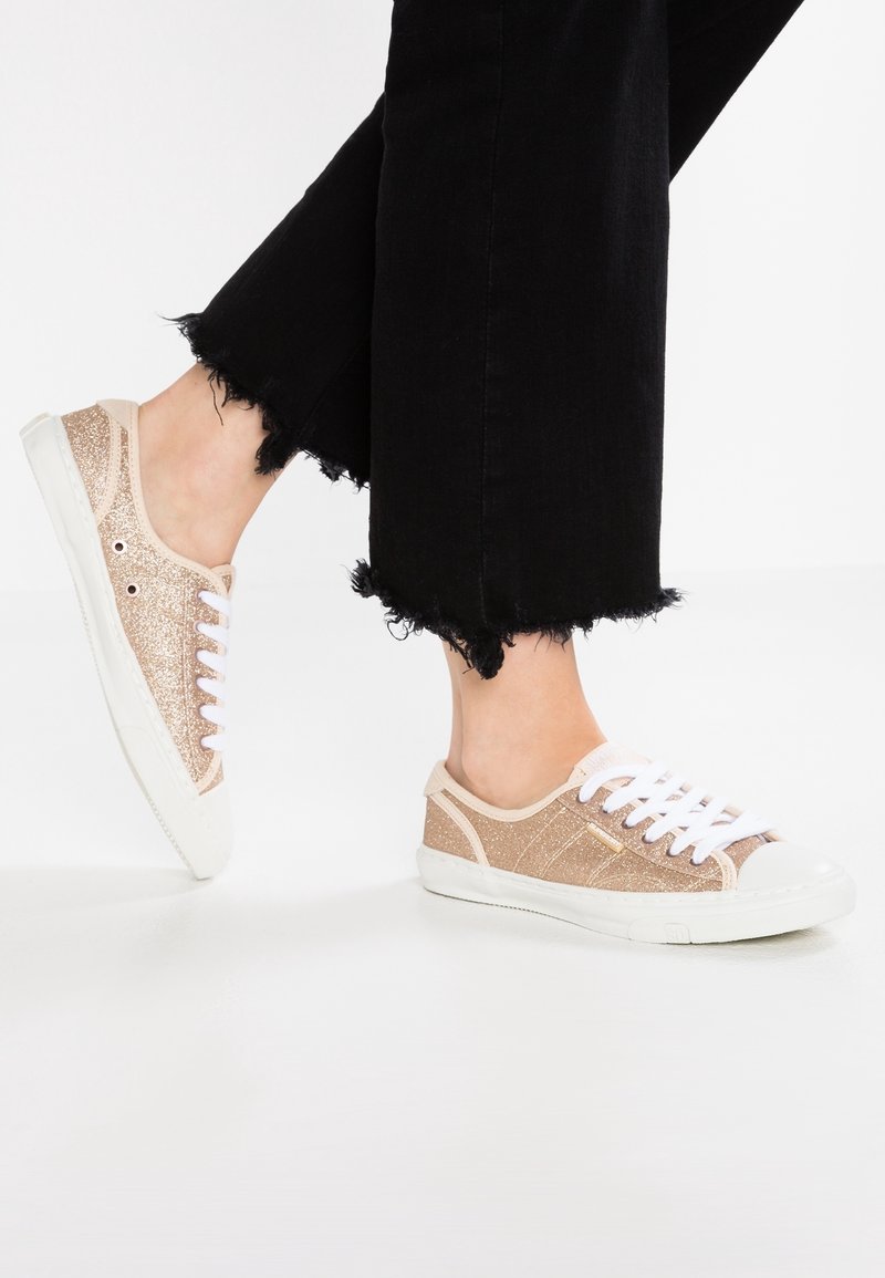 Zapatillas brillantes de color beige con cordones blancos y suelas de goma, con un upper texturizado y ribete blanco contrastante. Jeans negros con dobladillo deshilachado.