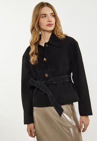 Schwarze Wildlederjacke mit einem Kragen-Design, Knopfschluss und Taillengürtel. Der Stoff hat eine glatte Textur und eine figurbetonte Silhouette.