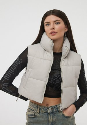 Gilet imbottito grigio chiaro con colletto alto, zip frontale e tasche a patta, indossato sopra una maglietta nera trasparente a maniche lunghe e jeans in denim.