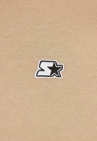 Logo bordado en blanco y negro que presenta una estrella y una "S" estilizada sobre un fondo de tela de punto beige.