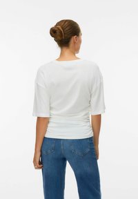 Haut blanc à manches courtes, avec une coupe décontractée et un détail froncé à l'arrière, associé à un jean bleu. Texture lisse et design simple.