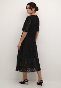 Black lace dress featuring short sleeves, a mid-length silhouette, and a floral pattern with a sheer underside. Paired with black high heels.