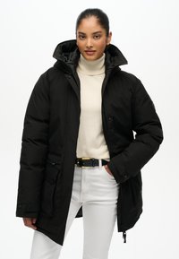 Czarna ocieplana parka z kapturem, zamkiem błyskawicznym i snapami, kieszeniami bocznymi oraz gładką fakturą, w połączeniu z białym golfem i spodniami.