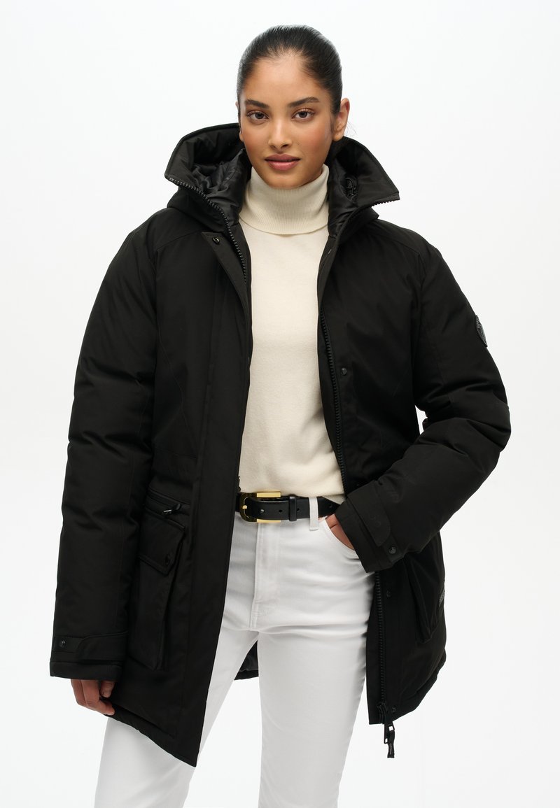 Czarna ocieplana parka z kapturem, zamkiem błyskawicznym i snapami, kieszeniami bocznymi oraz gładką fakturą, w połączeniu z białym golfem i spodniami.