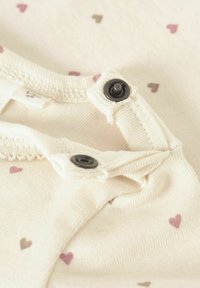 Vêtement pour bébé couleur crème avec des motifs de cœurs roses et bruns, doté de boutons-pression au niveau du col pour une fermeture facile.