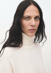 Maglione bianco a collo alto con texture a coste, caratterizzato da un collo alto. Il modello ha lunghi capelli scuri e uno sfondo neutro.
