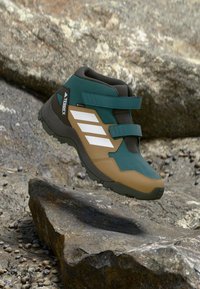 Grüner und beige halbhoher Wanderschuh mit einem strukturierten Obermaterial, weißen Akzenten und doppelten Klettverschlüssen; entworfen für anspruchsvolles Gelände.