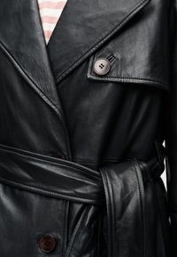 Κοντινή λήψη μαύρου δερμάτινου παλτού τρανς με μεγάλα κουμπιά, φαρδιούς γιακάδες και ζώνη δεμένη πάνω από ριγέ πουκάμισο.