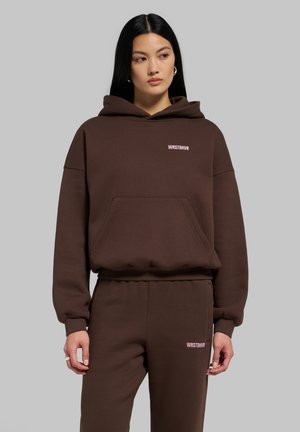 Femme portant un sweat à capuche marron et un pantalon de jogging assorti avec des logos blancs WRSTBHVR sur la poitrine et la cuisse, debout sur un fond gris.