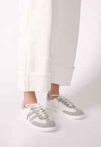 Sportliche Schuhe in Grau und Weiß, mit perforierten Einsätzen, Wildleder-Textur und einer weichen Gummisohle. Die Schnürsenkel sind weiß und durch Ösen gefädelt.