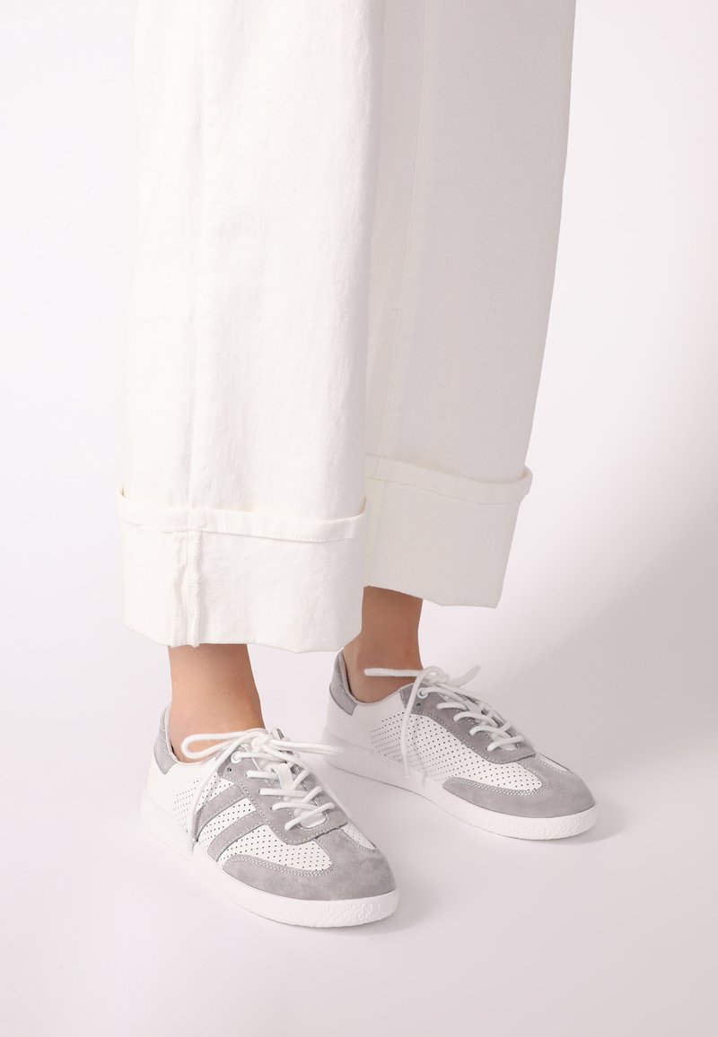 Sportliche Schuhe in Grau und Weiß, mit perforierten Einsätzen, Wildleder-Textur und einer weichen Gummisohle. Die Schnürsenkel sind weiß und durch Ösen gefädelt.