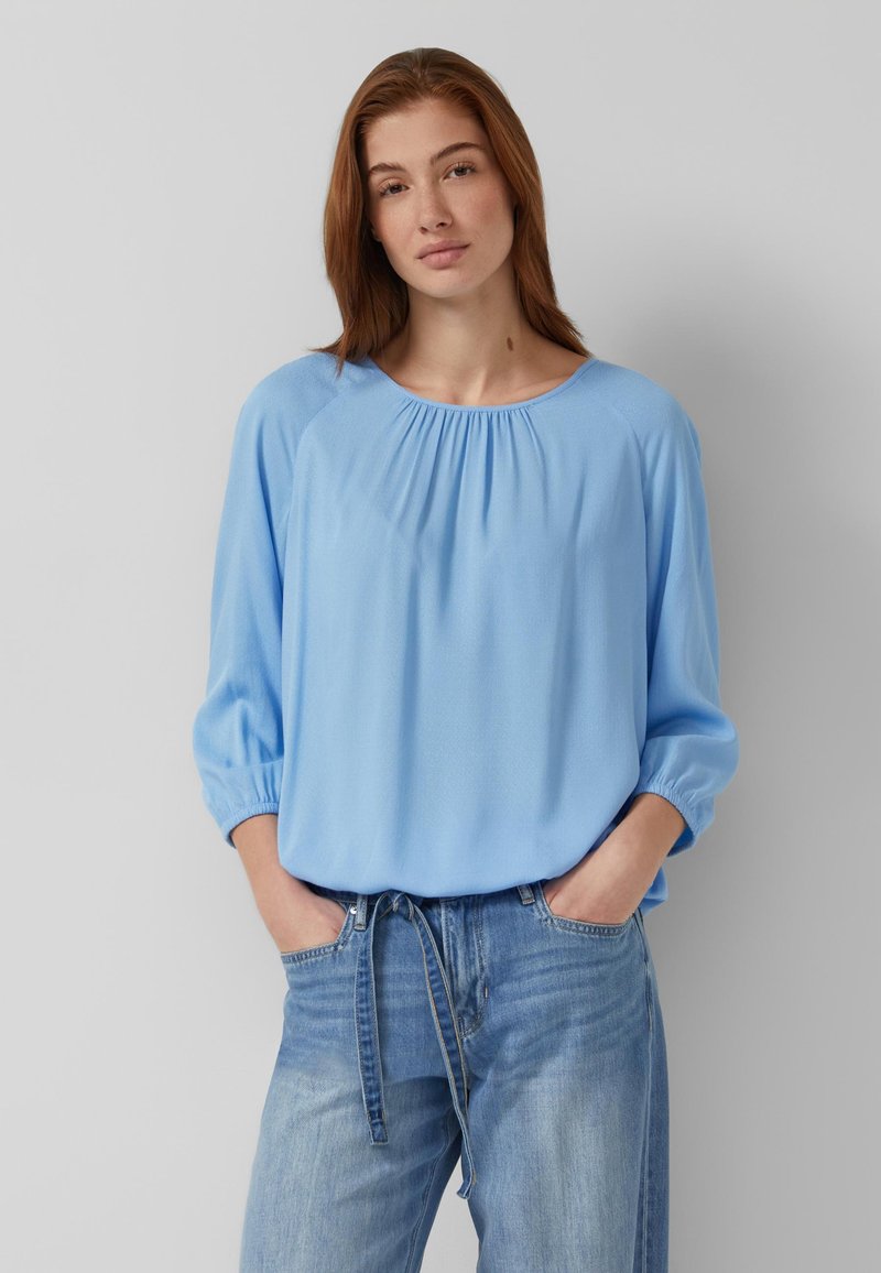 Frau mit roten Haaren, die eine hellblaue, lockere Bluse mit 3/4-Ärmeln und ausgewaschene blaue Jeans trägt, steht mit den Händen in den Taschen vor einer grauen Wand.