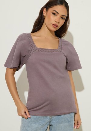 Frau trägt ein mauvefarbenes Top mit quadratischem Ausschnitt, Spitzenbesatz und kurzen Puffärmeln, kombiniert mit hellblauen Jeans, vor einem einfarbigen Hintergrund stehend.