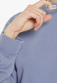 Hellelila Sweatshirt mit strukturiertem Finish, ausgestattet mit umgeschlagenen Ärmeln und einem silbernen Hardware-Detail am Kragen.