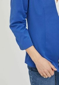 Blå skräddarsydd jacka med långa ärmar, strukturerad tyg och en kroppsnära silhuett. Matchas med denim, och visar en hand med ringar.