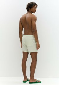 Világoszöld csíkozott fürdőnadrág, elasztikus derékkal és hátsó zsebbel, zöld papuccsal párosítva. Minimalista dizájn és textúra.