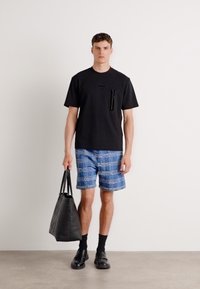 Svart kortermet T-skjorte med lommer, blå rutete shortser, svarte sokker, svarte sko og en svart totebag. Ensartet bakgrunn.