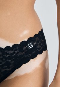 Svart blonderlingeri med bølgete kanter og et merke med teksten "HUGO." Designet har en semi-transparent tekstur.