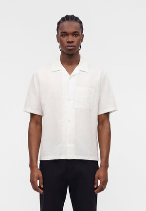 Mężczyzna z warkoczami, ubrany w białą koszulę z krótkim rękawem z kieszenią na piersi oraz czarne spodnie, stojący na jednolitym tle.