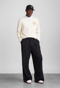 Kreemveida džemperis ar smalku dzīvnieku rakstu, apvienots ar melniem kvadrātiem svārkiem un baltām sporta kurpēm; melna cepure uz galvas.