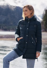 Donkergroene gewatteerde parka met een capuchon, zilverkleurige hardware, rits- en knoopsluitingen, en zijzakken. Draag met lichtblauwe spijkerbroek.