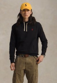 Zwarte hoodie met koordjes, rode logodetail, kaki cargobroek, meerkleurige gestreepte riem en een gele pet met logo. Vloeiende achtergrond.