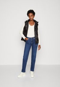 Gilet trapuntato nero con cappuccio, maglia a maniche lunghe crema, jeans blu a gamba dritta e sneakers bianche. Design semplice senza motivi visibili.
