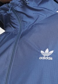 Marinblå lättviktjacka med dragkedja, texturerat tyg och vitt broderat Adidas-logotyp på bröstet.