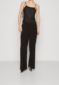 Zwarte mouwloze top met zilveren studs, gecombineerd met brede zwarte broeken. De outfit heeft een glanzende zilveren handtas en spitse zwarte hakken.